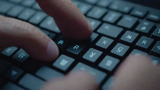 Manos escribiendo en un teclado retroiluminado frente al brillo de una pantalla.